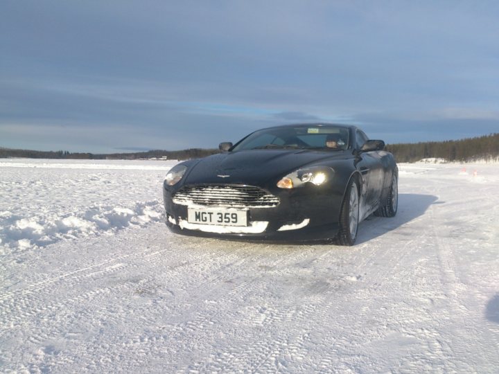 Aston Martin DB9 on Ice lake - Page 1 - Aston Martin - PistonHeads - The image features a black Aston Martin DB11 parked on a snowy track or runway. The car is positioned to the left of the frame and is facing the camera, with its headlights on. The ground is covered with a layer of light-colored snow that appears pristine and untouched, and the sky above is overcast with a blue tint. The car's peak is visible at the bottom of the image, and there are no other vehicles or people in the immediate vicinity, giving the scene a quiet, serene atmosphere.