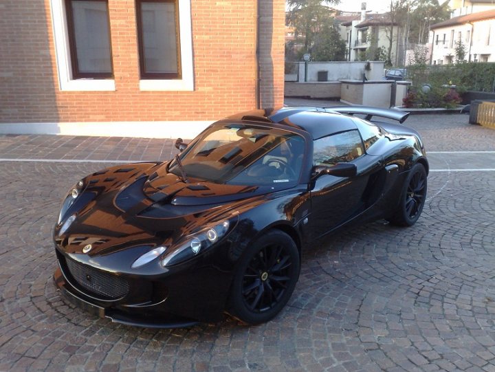 Story Pistonheads Drama Exige - The image depicts a modern, black sports car parked on a cobblestone street. The car is sleek, with a low profile and a large intake near the front. It features distinctive yellow headlights and black alloy wheels which suggest it might be a brand new model. The setting is urban, possibly outside a residential building, with clear skies and no other vehicles or pedestrians visible in the immediate vicinity. The contrast between the shiny black exterior of the car and the rough cobblestone surface adds a touch of sophistication to the scene. The parked car is the focal point of the image, taking up most of the frame and conveying a sense of luxury and speed.