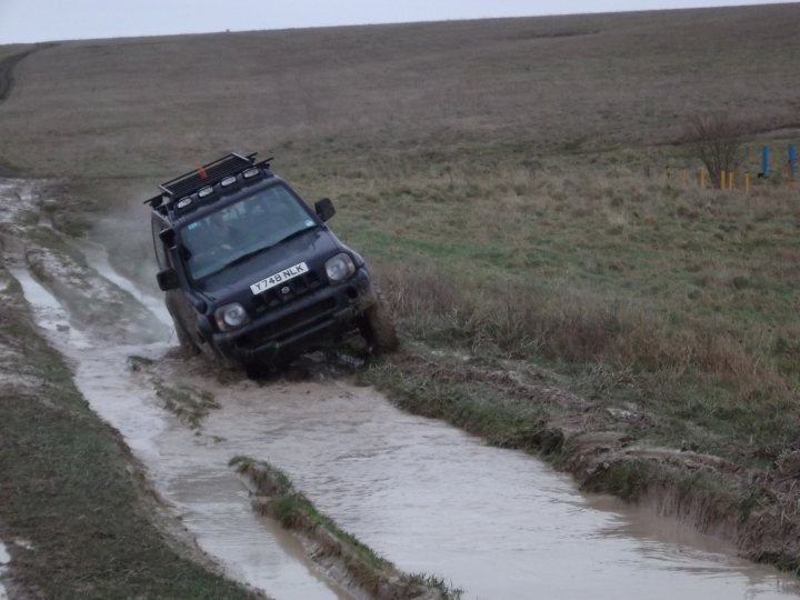 Pistonheads - The image shows a black SUV driving down a road where the road has become a muddy ditch due presumably to recent heavy rainfall. The car seems to be navigating its way through the muddy conditions with clear obstacles. The surroundings suggest a rural or undeveloped area, with fields on one side and what appears to be a fenced area on the other. The scene captures the aftermath of extreme weather, showing the car's capability to handle challenging terrain.