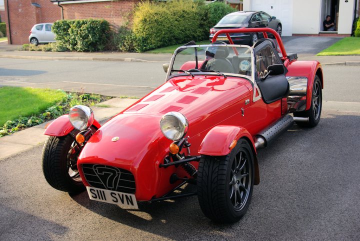 Metric chassis?? - Page 1 - Caterham - PistonHeads - The image is a photograph of a classic, open-top sports car, painted in a vibrant red color. The car features a prominent front bumper and two prominent circular headlights. The body of the car has horizontal slits and vents just behind the front wheels, contributing to its distinct design. Black wheels with well-defined spokes wrap around the tires on both sides. The backdrop is a suburban residential street with lawns, a driveway, and a partially visible house indicating a residential area. The sitting and sky regions of the image are dominated by the sky and clouds, suggesting it might be early morning or late afternoon.