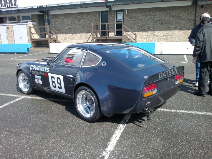 any 240z owners here? - Page 1 - Jap Chat - PistonHeads - The image showcases a vintage racing car, specifically a 1966 Datsun 200, parked in a racecourse parking lot. The car, painted in a rich black with white and red accents, is the center of attention and occupies a significant portion of the scene. On the side of the car, the number 89 is prominently displayed, suggesting its participation in racing events. The backdrop of brick buildings and a blue fence gives the setting a rustic charm. A few people are present in the image, likely spectators or crew members, adding a lively atmosphere to the scene.