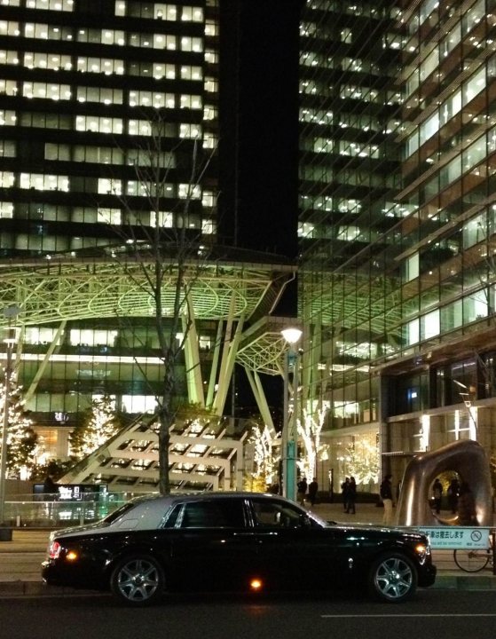 Spotted thread  - Page 1 - Rolls Royce & Bentley - PistonHeads - The image captures a city street at night, under the glow of high-rise buildings. The focus is on a sleek, black Cadillac, parked in front of a large, modern white building with a distinctive glass exterior and a unique curved roof. A cycle rack is also visible on the sidewalk next to the car. The atmosphere is enhanced by decorative Christmas lights adorning the building and street during the holiday season. The photo conveys a vibrant urban nightscape.