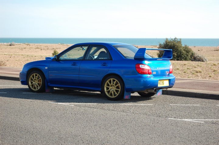 Prodrive Wrx Impreza Subaru Pistonheads Sti - The image shows a vibrant blue, two-door coupe parked on a beachside parking lot. The car features a large rear spoiler, a license plate, and gold-rimmed wheels, which reflect the color of the car. In the background, there's a serene beach with calm waves, providing a picturesque setting for the vehicle. The sky is clear and blue, suggesting a sunny day.