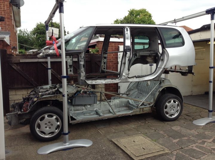 Lexus V8 with NOS in a Renault Espace - yeah lets do it !  - Page 12 - Readers' Cars - PistonHeads - The image shows an old, heavily-damaged white van parked outside on a gray day. The van appears to be in a state of disrepair with visible signs of wear, such as rust and paint coming off the body. The interior has been stripped out, revealing old upholstery and mechanics. The van is propped open by various support structures indicating it might need repair or is in the process of being restores. There is a brick wall visible in the background and a grassy area behind the van. The scene seems to be filmed with a handheld camera, as suggested by the grainy quality of the video. The image evokes a sense of the van having been involved in a severe accident or other destructive incident.