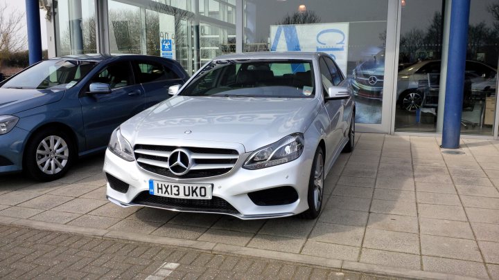 Anyone taken delivery of a new shape e class sport - Page 2 - Mercedes - PistonHeads - The image shows a silver Mercedes-Benz car parked in a dealership lot, adjacent to a blue hatchback. The dealership has a modern architecture, with large glass windows that reflect the cars and the blue color of one of the pillars. In the background, another Mercedes-Benz vehicle is visible, adding to the commercial atmosphere of the setting. The sale sign "HX13 UKO" is visible on the silver car, indicating that the car is from the United Kingdom. The sky outside the dealership is gray, suggesting an overcast set of weather conditions.