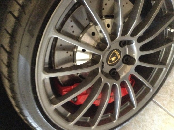 Advice please - Page 1 - Gallardo/Huracan - PistonHeads - The image shows a close-up of a car's wheel as seen from the side. The rim of the wheel is silver and features a traditional five-point logo, which is emblematic of a certain luxury car manufacturer. The hubcap is red with a design that appears to have a black center. The car's brake rotor is visible, suggesting that the wheel is either off a hub or has a transparent central spacer that allows a clear view of the rotor. The ground on which the wheel is placed is a light color, providing a neutral background that contrasts with the metallic and vibrant colors of the wheel.