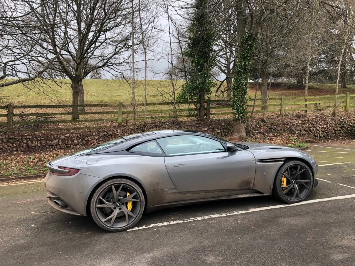 DB11 / DBSS tyre choice - Page 1 - Aston Martin - PistonHeads - The image shows a gray Aston Martin DB11 parked in what appears to be an outdoor parking area. The car is sleek, with the distinctive design and proportions typical of high-end sports cars. It features unique wheels and a yellow license plate. In the background, there's a fence and some trees, suggesting this might be a private estate or a dedicated parking space. There are no people visible in the image.