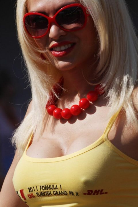 Grid Girls/Pit Babes (Vol 3) - The image features a person with a fair complexion, wearing a vibrant yellow crop top and polarizing red sunglasses. The individual's hair is blonde and styled, and they sport a red bead necklace. The backdrop of the image is blurred, keeping the focus on the person in the foreground. This portrayal suggests a casual, outdoor setting, possibly during the day.