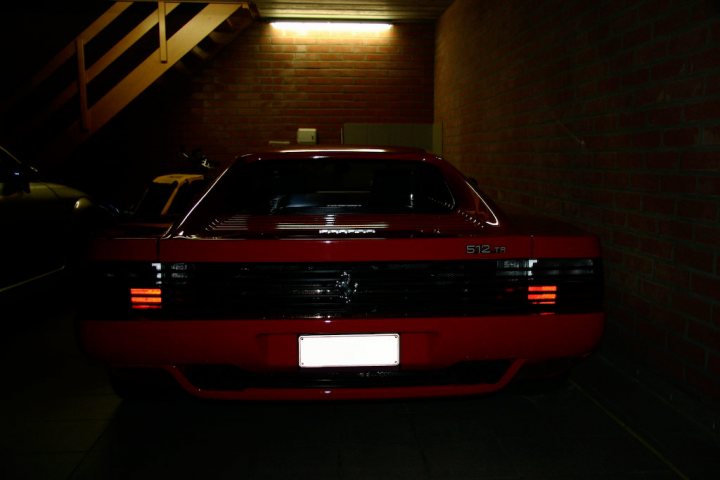 My 512TR - Page 1 - Ferrari V12 - PistonHeads - In the image, we see a side view of a parked car, captured at night. The car is situated in a dimly lit garage or staircase area, suggesting it might be a public space or a shared parking area. The vehicle is a red sedan, as indicated by its license plate which reads "512T". Its windshield is clean, reflecting the ambient light of the surroundings.