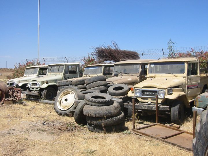 Classics left to die/rotting pics - Page 80 - Classic Cars and Yesterday's Heroes - PistonHeads - The image shows an assortment of vehicles parked in an open area on what appears to be a sunny day. There are four trucks of varying sizes, arranged in a somewhat haphazard manner. The trucks have a rugged appearance and are not in pristine condition. There are also several tires scattered around the scene. The vehicles appear to be older models, suggesting they might not be in active use. In the background, there's a wire fence, indicating that the area could be a private property or a commercial or industrial facility.